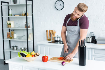 smiling bearded man in apron talking by smartphone and cutting vegetables in kitchen