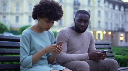 Girlfriend and boyfriend chatting in their smartphones, sitting on bench, ignore