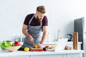 handsome bearded man in apron using laptop and cooking vegetable salad