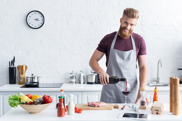 handsome bearded young man in apron pouring wine and smiling at camera while cooking in kitchen - Powered by Adobe