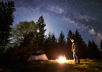 Camping site in mountain valley at night. Male hiker standing in front of tourist tent at burning campfire, pointing at night blue starry sky with Milky way, full moon. Outdoor activity concept