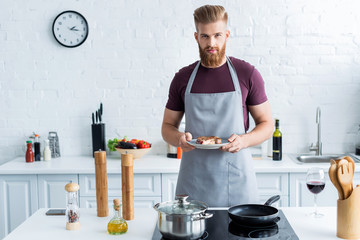 handsome bearded young man in apron holding delicious beef steak on plate and looking at camera