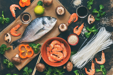 Ingredients for Asian dishes - raw Dorado fish, shrimp, rice noodles,mushrooms, vegetables, dark background, top view. Flat lay, toned photo.