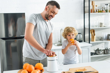 father making orange juice by squeezer while his little son eating orange at kitchen