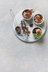 Three servings of chocolate ice cream, decorated with chocolate and mint in white bowls on round stone board. Close-up, top view on gray concrete background