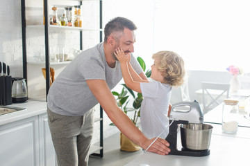 side view of little boy touching smiling father cheeks near tabletop at kitchen