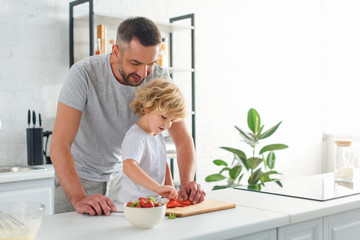 Obraz na płótnie Canvas man standing with son while he taking strawberry from cutting board at kitchen