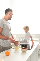 side view of father whisking dough in bowl and talking to son while he sitting on tabletop at kitchen