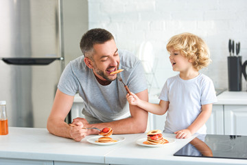 smiling little boy feeding father by pancake at kitchen