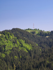 A high telecommunication tower with antennas, painted aviation red and white,  stands on the top of mountain covered with forest. Taken on a sunny day. Background image of nature and technology.