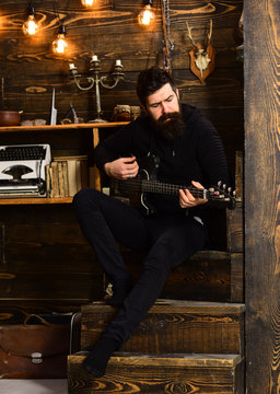 Boost your skills. Guy in cozy warm atmosphere learn new song chords. Man bearded musician enjoy evening rehearsing performance at home, wooden background. Man with beard holds black electric guitar