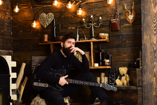 Man bearded musician enjoy evening with bass guitar, wooden background. Guy in cozy warm atmosphere play relaxing soul music. Favourite activity. Man with beard holds black electric guitar