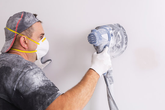 Plasterer wearing dust mask polishes a wall with sanding machine . House renovation concept.