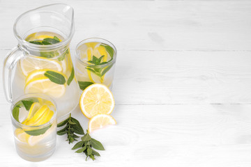 Homemade lemonade with lemon and mint in a glass jug and a glass next to fresh lemon on a white wooden background