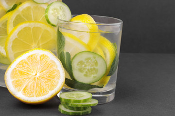 Refreshing water with lemon and cucumber and mint in a glass cup and in a jug next to half a fresh lemon and cucumber slices on a black background