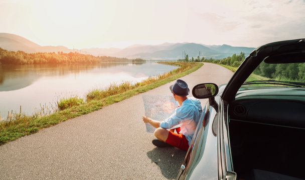 Man reads roads map sitting near his cabriolet on picturesque mountain road
