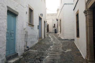 Fototapeta na wymiar A view of a narrow street with arch and wooden windows and doors with white wall stone architecture of the island Patmos, Greece 