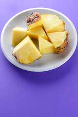 Pieces of pineapple on a white plate, chopped fresh pineapple on a violet background, salad of tropical fruits for breakfast, vegetarian food, copy space