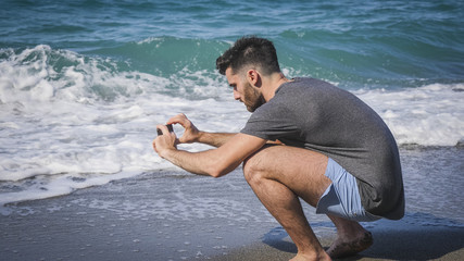 Full body shot of a handsome young man using cell phone to take photo, crouching on a beach, wearing boxer shorts and dark t-shirt