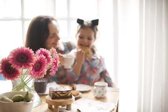 A Happy Family Is Having Breakfast At Home By The Window At The Table. Flowers And Coffee. Mom And Daughter. Copy Space.