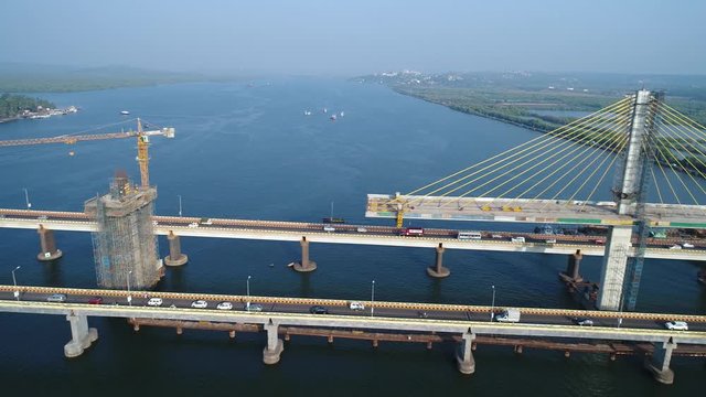 The camera moves over a large automobile bridge on which cars drive, past the cranes that are building new sections of the cable-stayed bridge on Mandovi river in Goa, India. Aerial view.
