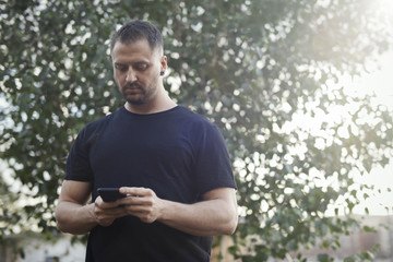 Fototapeta premium Young man typing text message on mobile phone outdoors in the park.
