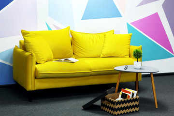 Comfortable yellow sofa against colored wall with geometrical figure.Box with books near soafa and coffee table with green plant. Relaxing zone.Copy space