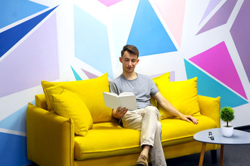 Young man sitting on comfortable yellow sofa and read book. Against colored wall with geometrical figure.Coffee table with green plant near sofa. Relaxing zone.Copy space