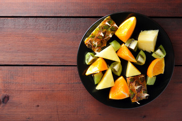 Fruit salad for vegans, pieces of pineapple, oranges and kiwi on a black plate, tropical fruits on wooden boards, citrus salad top view, copy space