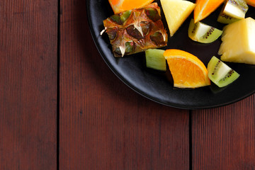 Fruit salad for vegans, pieces of pineapple, oranges and kiwi on a black plate, tropical fruits on wooden boards, citrus salad top view, copy space