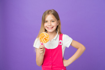 Smiling child with bright lollipop. Girl with long blond hair in pink outfit isolated on purple background. Sweet tooth with happy face holding big round candy, dessert time concept
