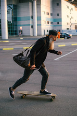 Stylish handsome middle-aged man with long gray beard riding on longboard during sunset  on urban background