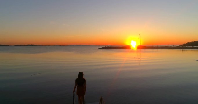Woman looking at the sea, C4K aerial backward view over a girl enjoying the sunset, at a wharf, on a beach at the baltic sea, on a sunny summer evening dusk, Hanko, Finland