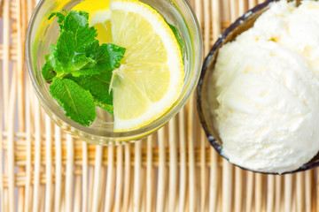 Fresh Cool Infused Detox Water with Ripe Organic Sliced Lemons Mint in Frosty Glass Ice Cream in Coconut Bowl on Rattan Table. Healthy Drink Dessert Summer Refreshment. Top View Copy Space