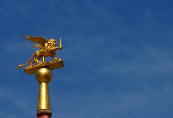 Fototapeta na wymiar Saint Mark Winged Lion golden statuette wielding a sword against blue sky, a symbol of the Old Venice Republic at war (with copy space)
