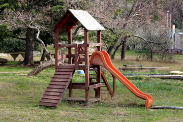 Fototapeta na wymiar Outdoor wooden public playground equipment with climbing steps and plastic slide surrounded with winter vegetation and uncut grass