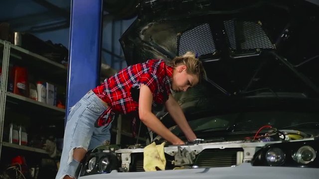 Sexy cute blonde girl in ripped jeans and red plaid shirt repairs car and to screws detail of engine with help wrench under hood of car in service station