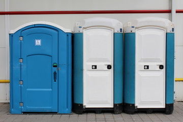Three portable blue and white ecological toilets in a row for men, woman and disabled persons in front of grey building and on concrete tiles
