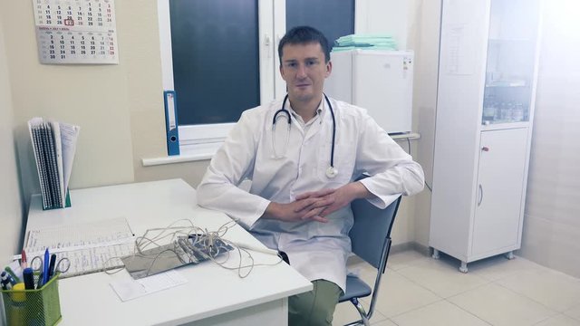 Young doctor with stethoscope sits on a chair by the table in his office