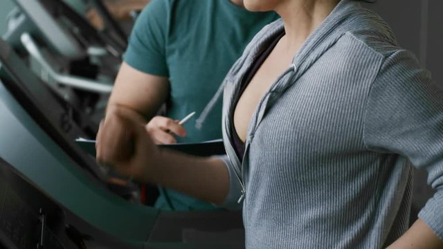 Tilt down shot of concentrated woman exercising on treadmill at the gym, coach taking notes and adjusting speed
