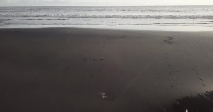 Fit surfer lady in sexy bikini and short shorts with white surfing board running on a black beach at sunrise. Modern lifestyle. Sport camp and extreme swim on summer vacation. Aerial drone footage.
