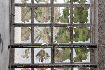 Small windows with broken glass, rusted frames and tall pine trees in background surrounded with dilapidated walls in abandoned building