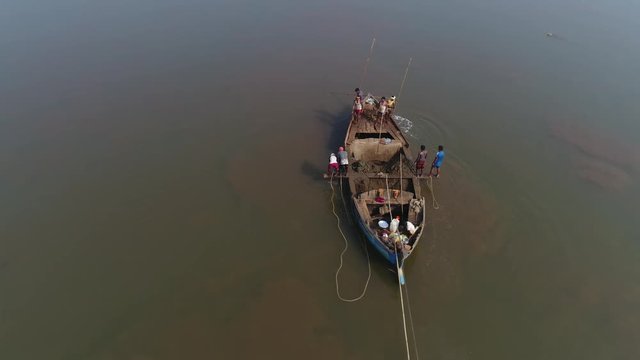 Aerial view. Flying around a small boat on which several Indian workers extract sand from the bottom of the muddy river by hand, with the help of sticks, buckets and ropes. 