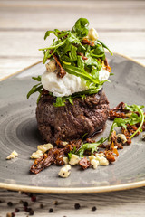 a large piece of juicy steak with egg poached and rucola salad with goat cheese in olive oil, lies on a plate. On the table is a glass of red wine. concept of a beautiful dish serving for a restaurant