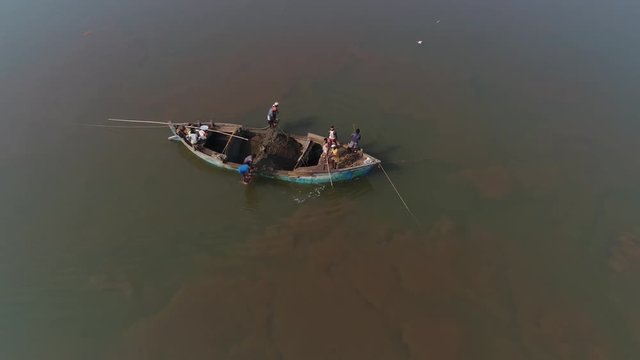 People on the small boat take out the soil from the bottom of the river with the help of sticks of buckets and ropes. Aerial view.