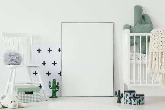 Grey pompom placed on white chair standing in bright baby bedroom interior with cactus shaped decor and empty poster on the floor. Put your picture here