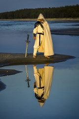 Portrait of assassin in white costume with the sword at the sea. He is posing near water with reflections on it. Sunset time. Isolated. - 212038108