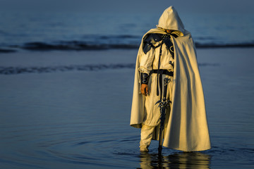 Portrait of assassin in white costume with the sword at the sea. He is posing near water with reflections on it. Sunset time. Isolated. - 212038169