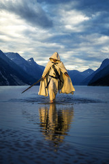 Portrait of assassin in white costume with the sword at the sea. He is posing near water with reflections on it. Sunset time. Isolated. - 212038175
