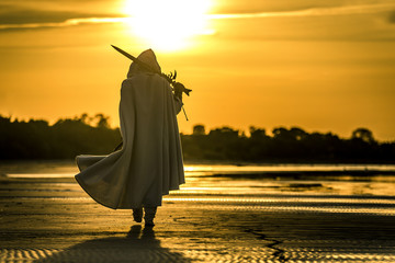 Portrait of assassin in white costume with the sword at the sea. He is posing at the sea during beautiful sunset. Yellow, soft light. - 212038730